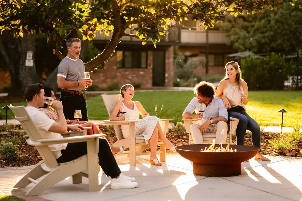 Group of friends relaxing by a firepit in a garden laughing and drinking wine.