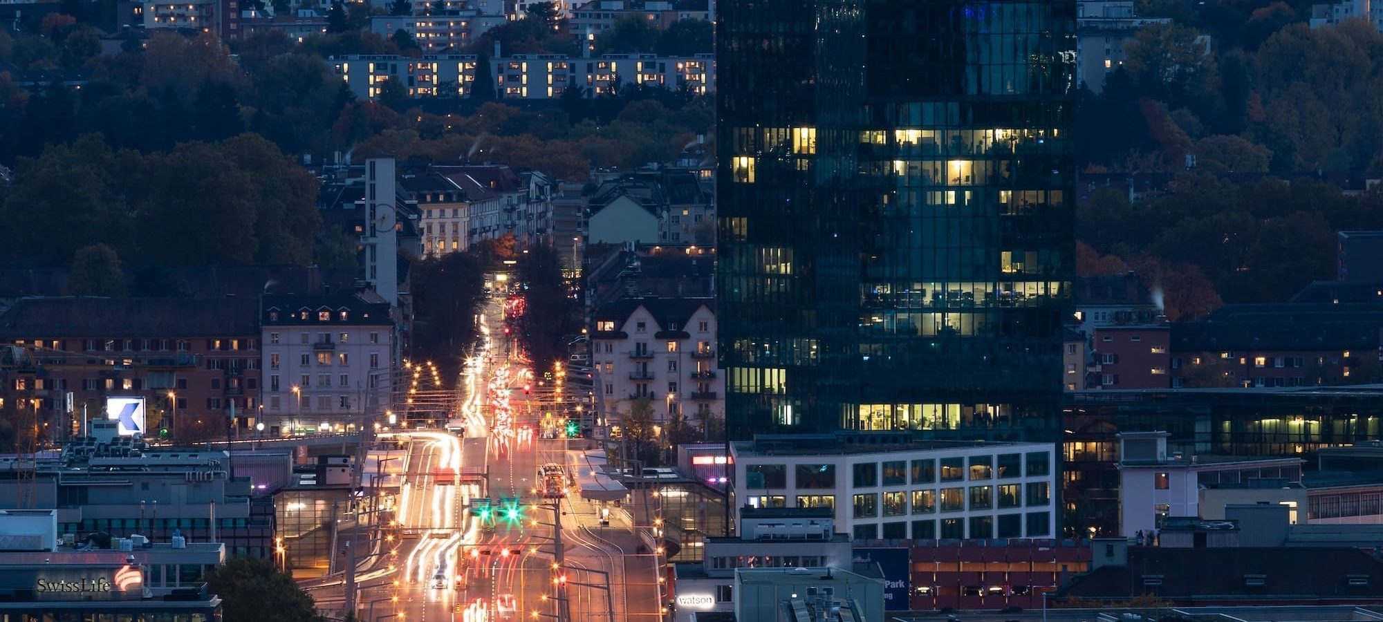 A tall, modern glass skyscraper stands illuminated in a city at dusk, with blurred car lights trailing along a busy road leading toward the building. Surrounding structures and hills are visible in the background.