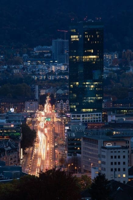 A tall, modern glass skyscraper stands illuminated in a city at dusk, with blurred car lights trailing along a busy road leading toward the building. Surrounding structures and hills are visible in the background.