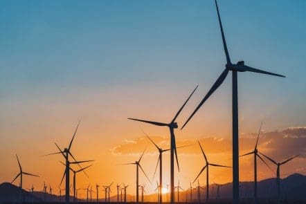 Wind turbines stand in a field with mountains in the background, silhouetted against a colorful sunset sky with orange and blue hues.