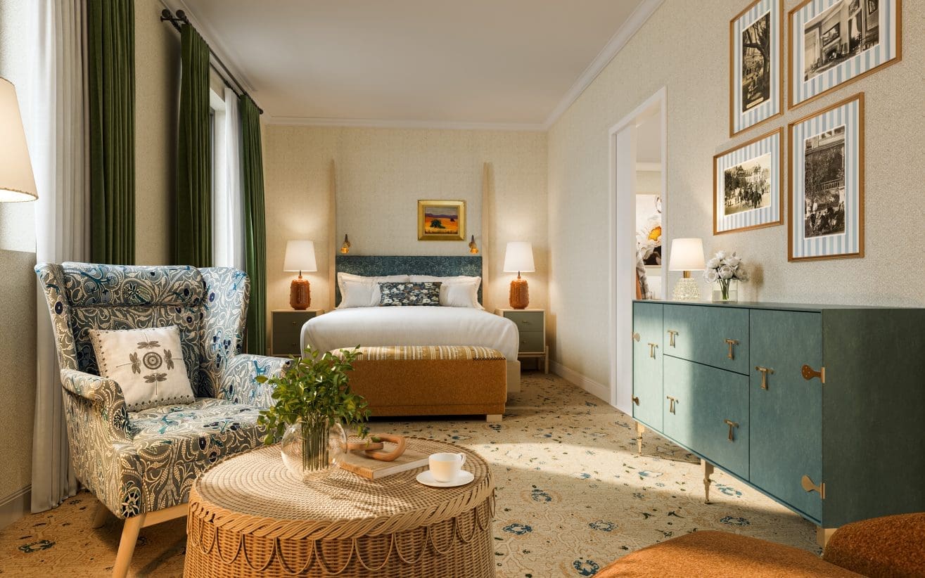 A cozy, well-lit hotel room features a double bed with a green headboard, a patterned armchair, a wicker table with a plant and cup, framed art on the walls, and a green dresser with decor.