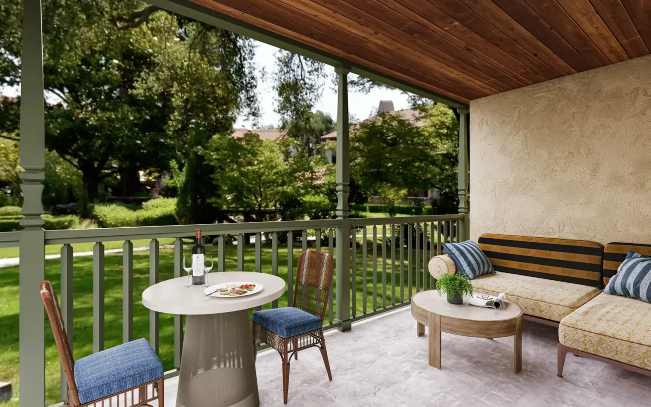 A covered balcony with a round table set for two, wine, and food. There’s a cushioned outdoor sofa and coffee table, overlooking a lush green garden with trees and sunlight filtering through.