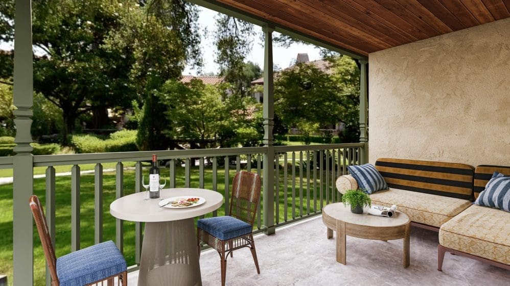 A cozy covered patio with a table set for two, two chairs, a small sofa with cushions, and a coffee table, overlooking a lush green garden with trees and manicured lawns.