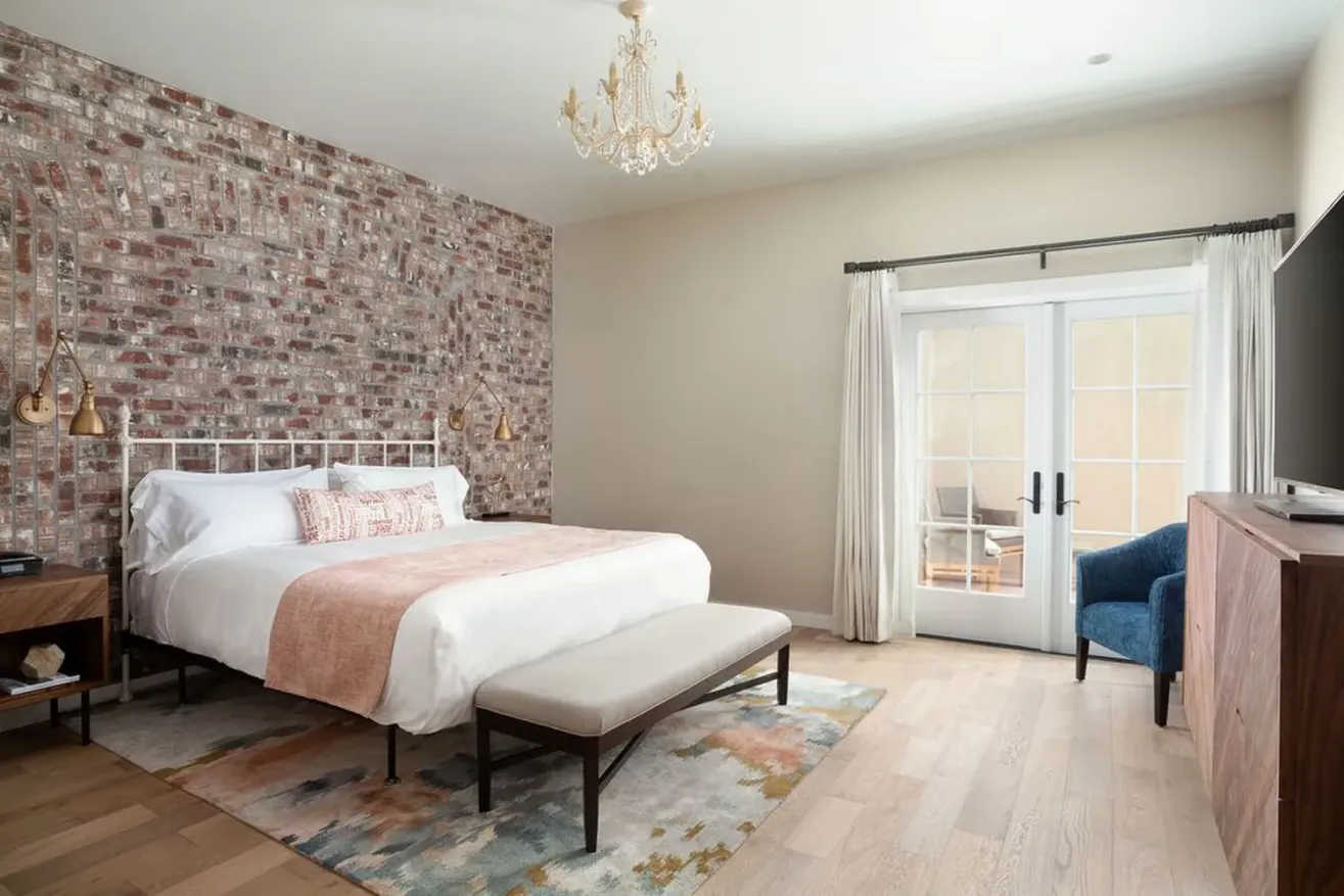 A modern bedroom with a brick accent wall, white bed with peach accents, wooden furniture, a chandelier, a blue armchair, French doors with curtains, and a colorful rug on light wood floors.