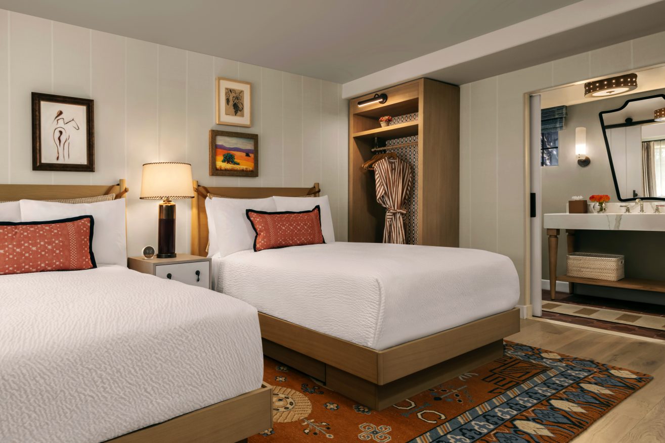 A hotel room with two double beds, white bedding, patterned pillows, a nightstand with a lamp, open closet with hanging robe, artwork on the walls, and a bathroom visible through an open door.