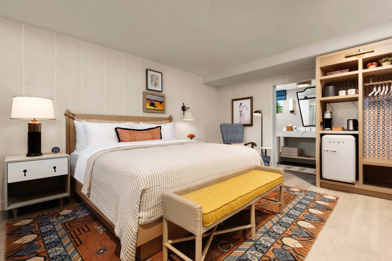 A cozy hotel room with a large bed, white bedding, a yellow bench at the foot, a nightstand with a lamp, an open wardrobe, a small fridge, a rug, and a bathroom area visible in the background.