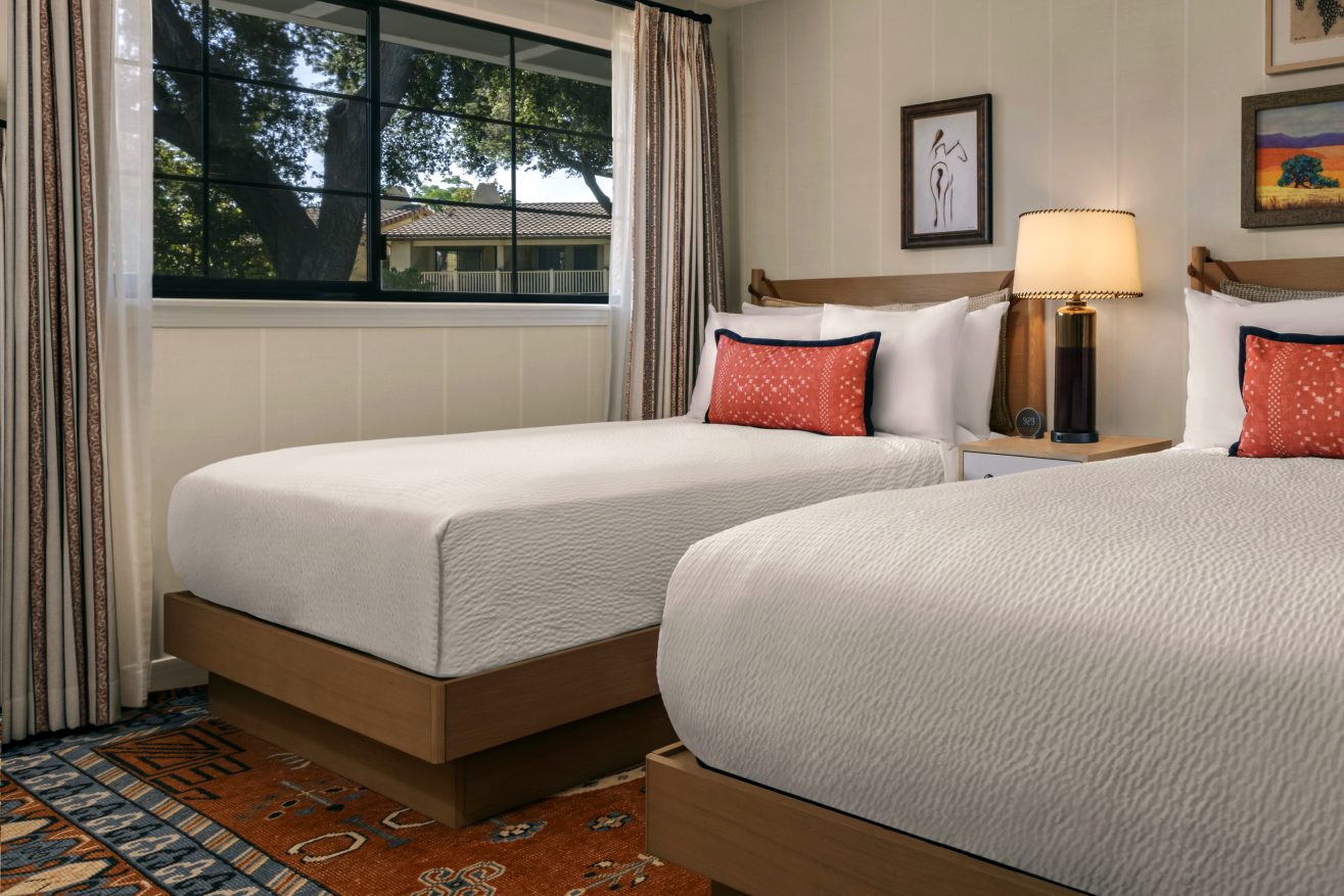A cozy bedroom with two neatly made twin beds, white bedding, orange accent pillows, a bedside table with a lamp, framed art on the walls, a colorful rug, and a window showing trees outside.