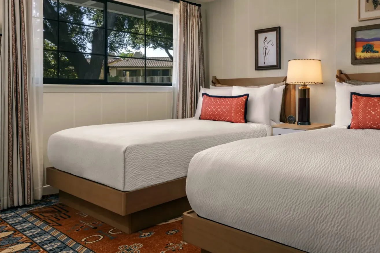 A cozy bedroom with two neatly made twin beds, white bedding, orange accent pillows, a bedside table with a lamp, framed art on the walls, a colorful rug, and a window showing trees outside.
