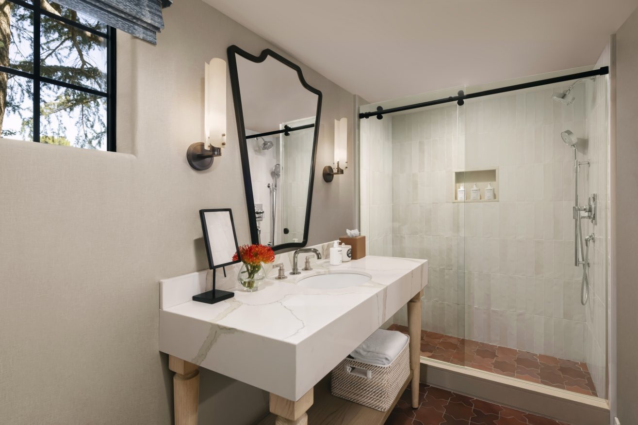 A modern bathroom with a large white marble sink, a decorative mirror, wall sconces, and a glass-enclosed shower. A tray with flowers and toiletries sits on the counter, with towels stored on a shelf below.
