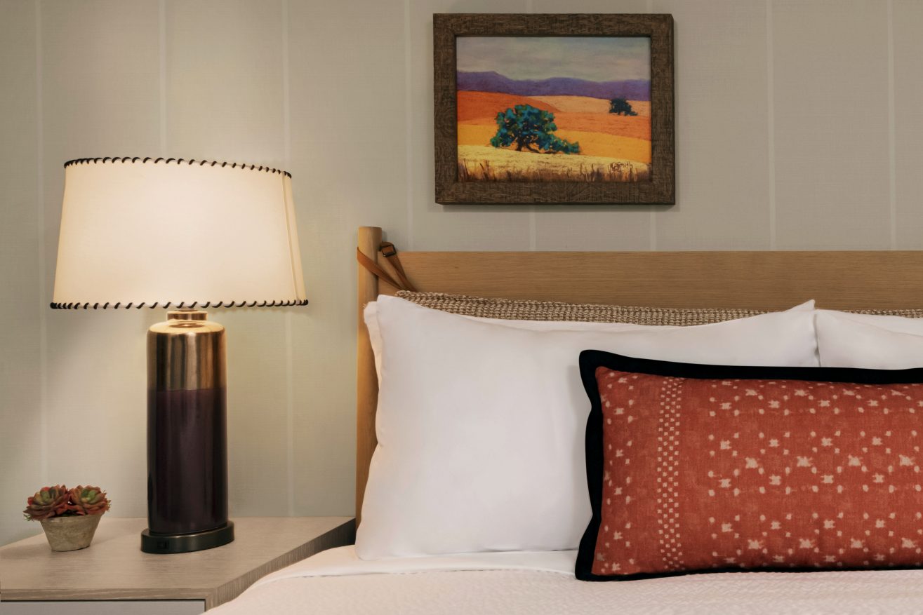 A cozy bedroom scene with a bed, white pillows, a red patterned cushion, a bedside table with a modern lamp, a small succulent plant, and a painting of a landscape with orange fields on the wall.