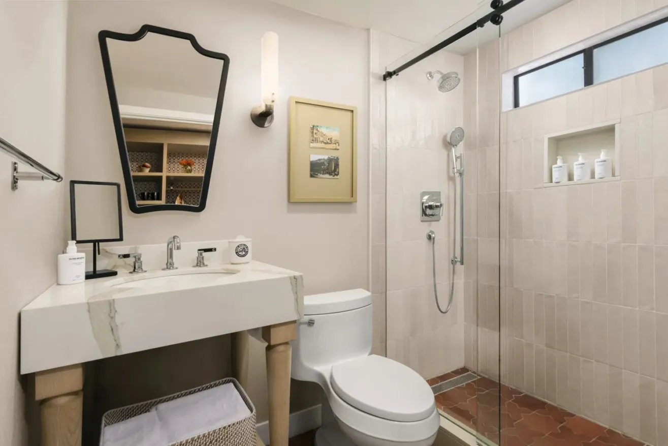 A modern bathroom with a white sink and marble countertop, a toilet, a glass shower with beige tiles, a wall mirror, a small wall shelf with toiletries, and framed artwork on the wall.