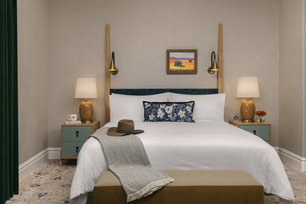 A neatly made bed with white linens, a patterned pillow, and a gray blanket with a hat on top. There are two nightstands with lamps, a mug, and flowers. A small landscape painting hangs above the bed.