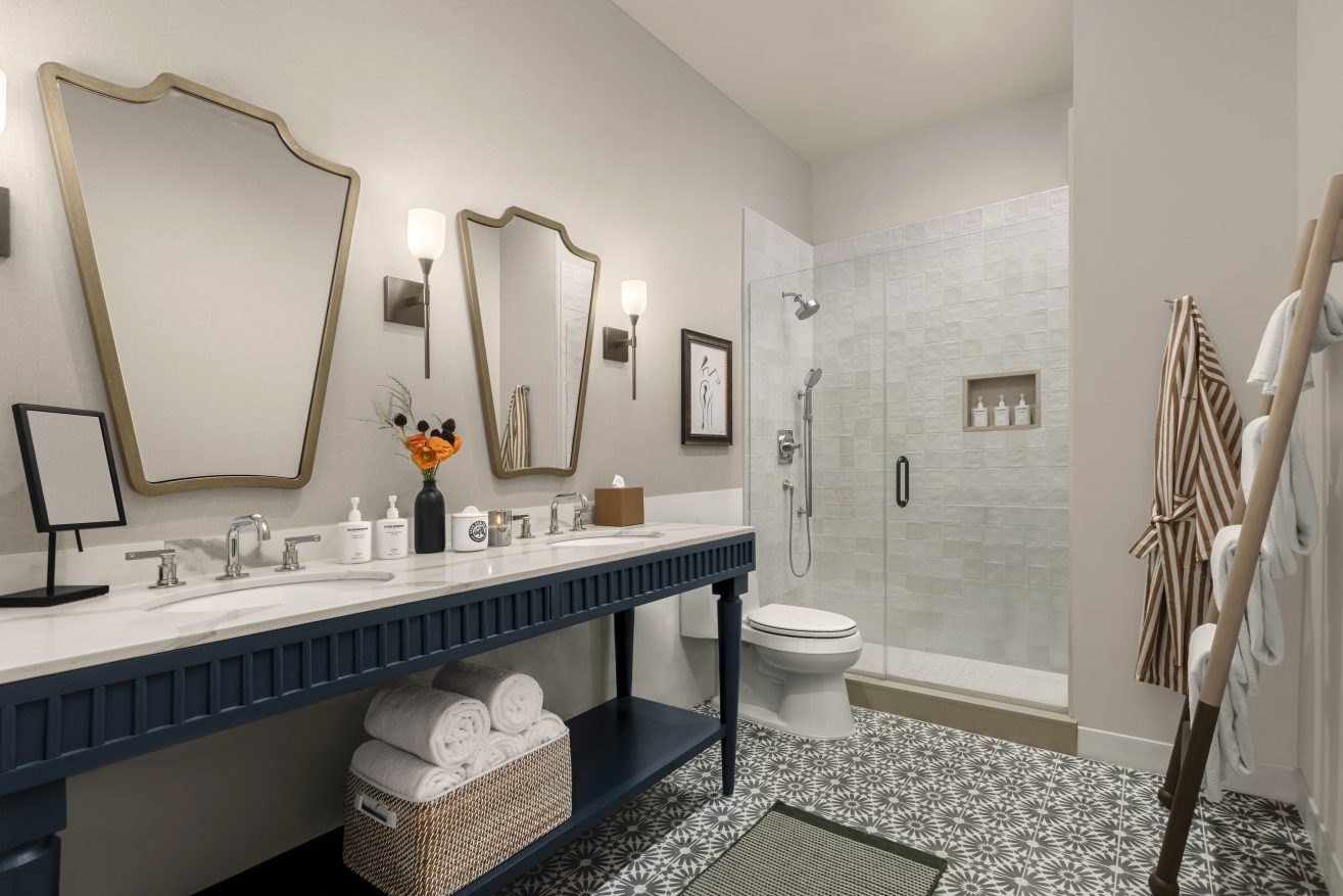 Modern bathroom with a double sink vanity, two mirrors, wall sconces, stacked towels, a glass-enclosed shower, patterned floor tiles, and a robe hanging on a wooden rack.