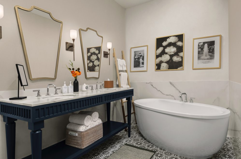 A modern bathroom with a blue double-sink vanity, two ornate mirrors, a freestanding bathtub, three framed pictures on the wall, and rolled white towels neatly stored below the counter.