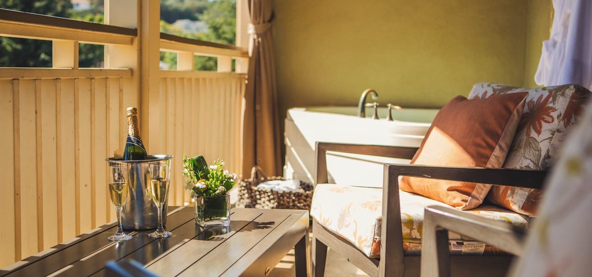 A sunlit balcony with cushioned chairs, a wooden table holding a wine bucket with champagne and glasses, a potted plant, and a bathtub in the corner. Trees and greenery are visible outside.