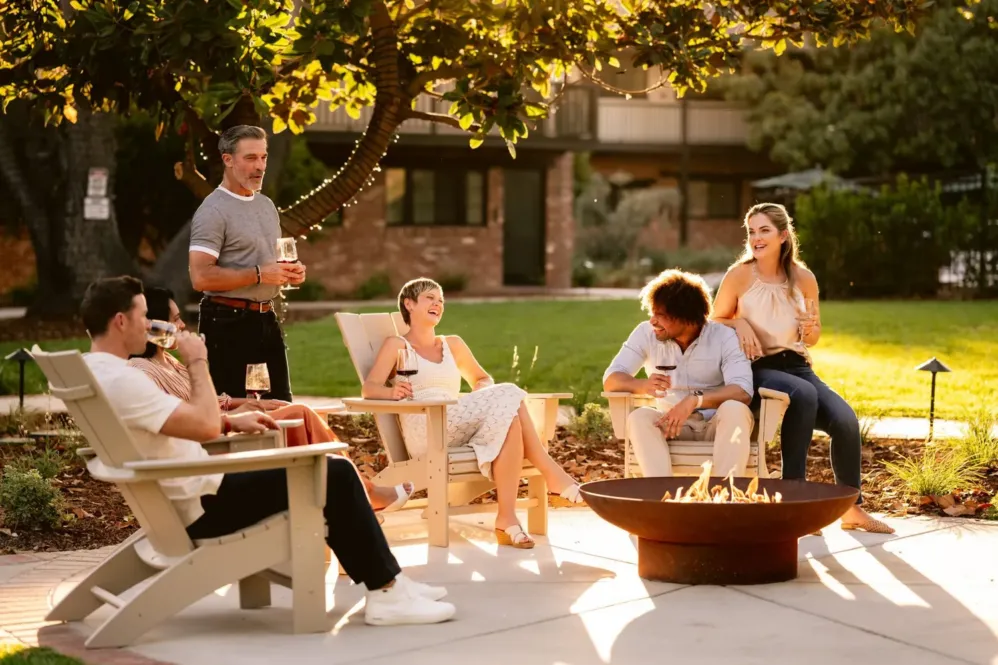 Group of friends relaxing by a firepit in a garden laughing and drinking wine.