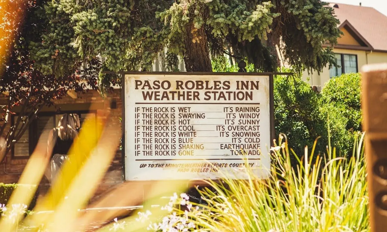 Rustic wooden sign reading "Paso Robles Inn Weather Station," surrounded by lush greenery, with a charming brick pathway in the background.
