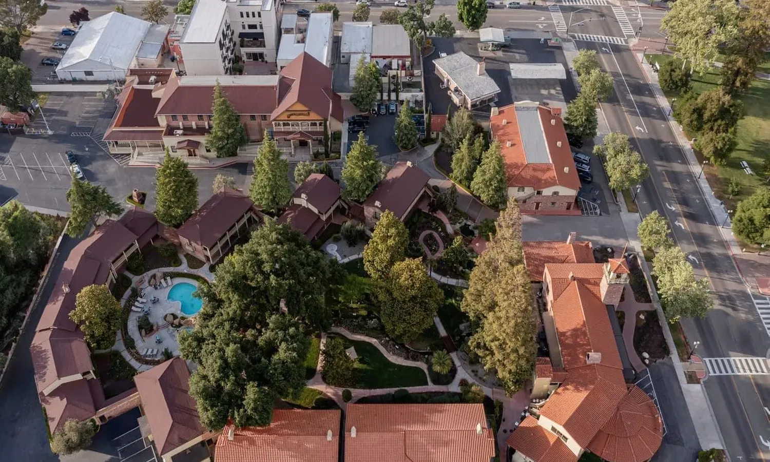 Aerial view of a complex with red-roofed buildings, central gardens, winding paths, and a swimming pool; surrounded by roads, parked cars, and nearby trees.