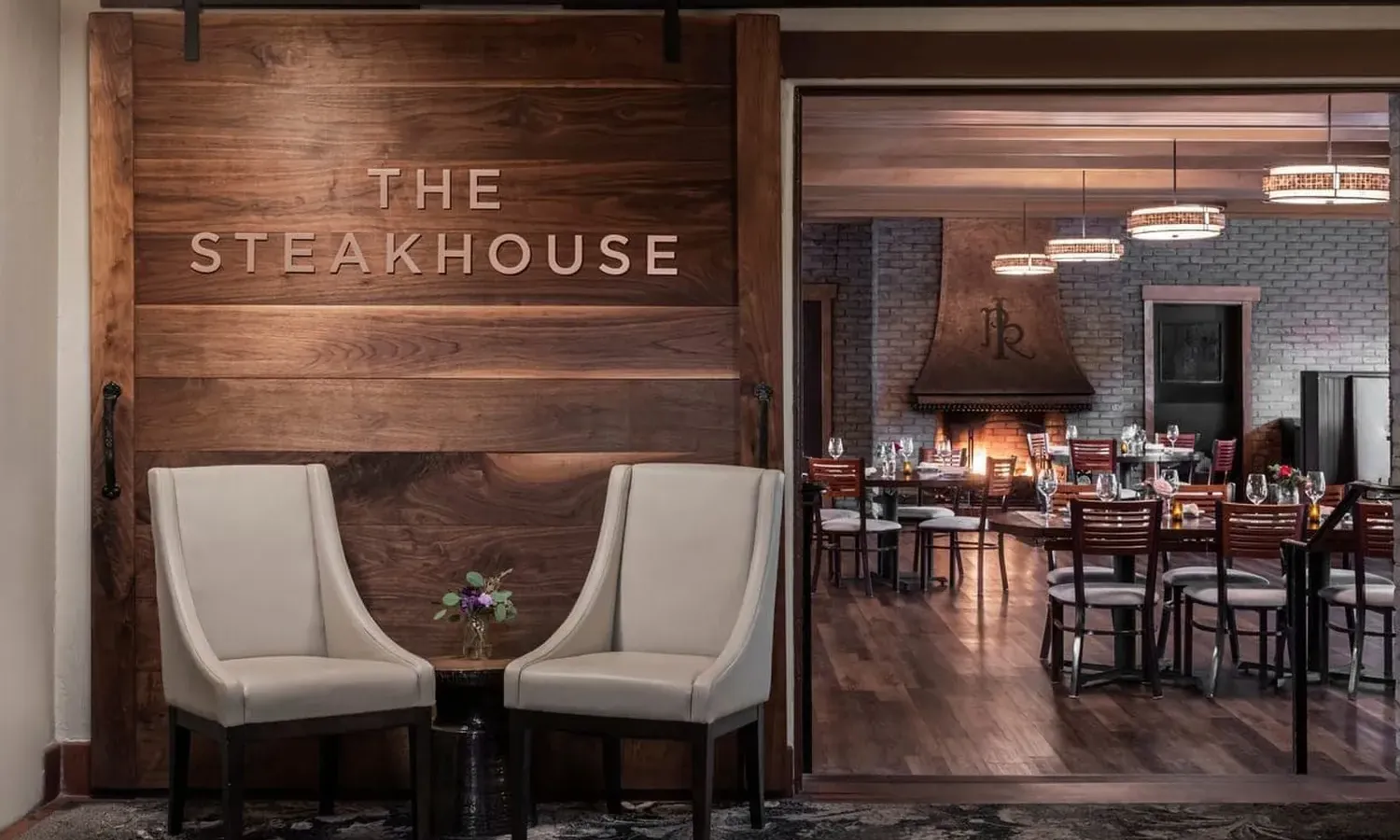 Two beige chairs and a small table with flowers sit in front of a large wooden door labeled The Steakhouse, with a view into a modern, warmly lit dining area in the background.