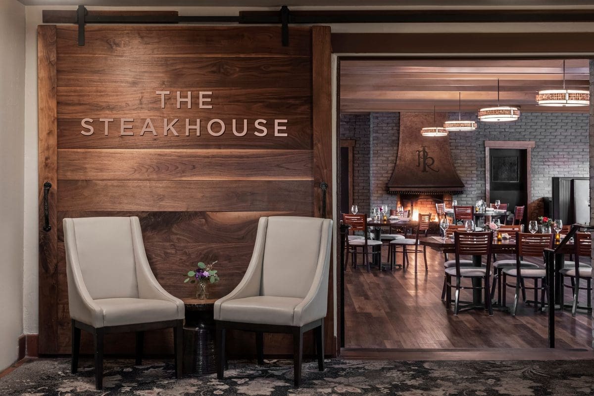 Two beige chairs and a small table with flowers sit in front of a large wooden door labeled The Steakhouse, with a view into a modern, warmly lit dining area in the background.