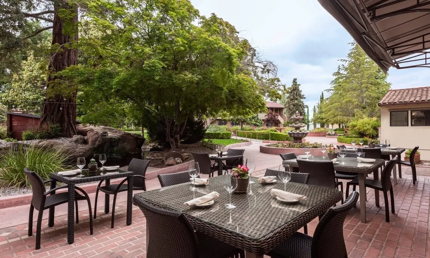 A cozy outdoor lounge area with a fire pit, surrounded by comfortable seating, lush plants, and natural daylight.