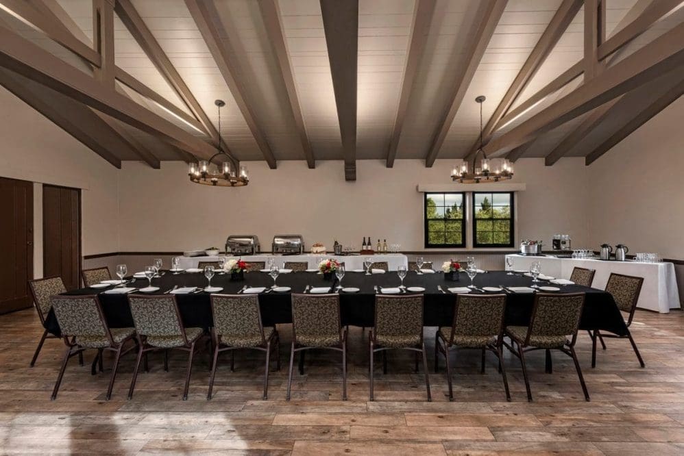 A large dining room with a long rectangular table set for a formal meal, surrounded by chairs. Buffet tables with food, drinks, and tableware are along the back wall. The room has wooden floors, exposed beams, and windows.