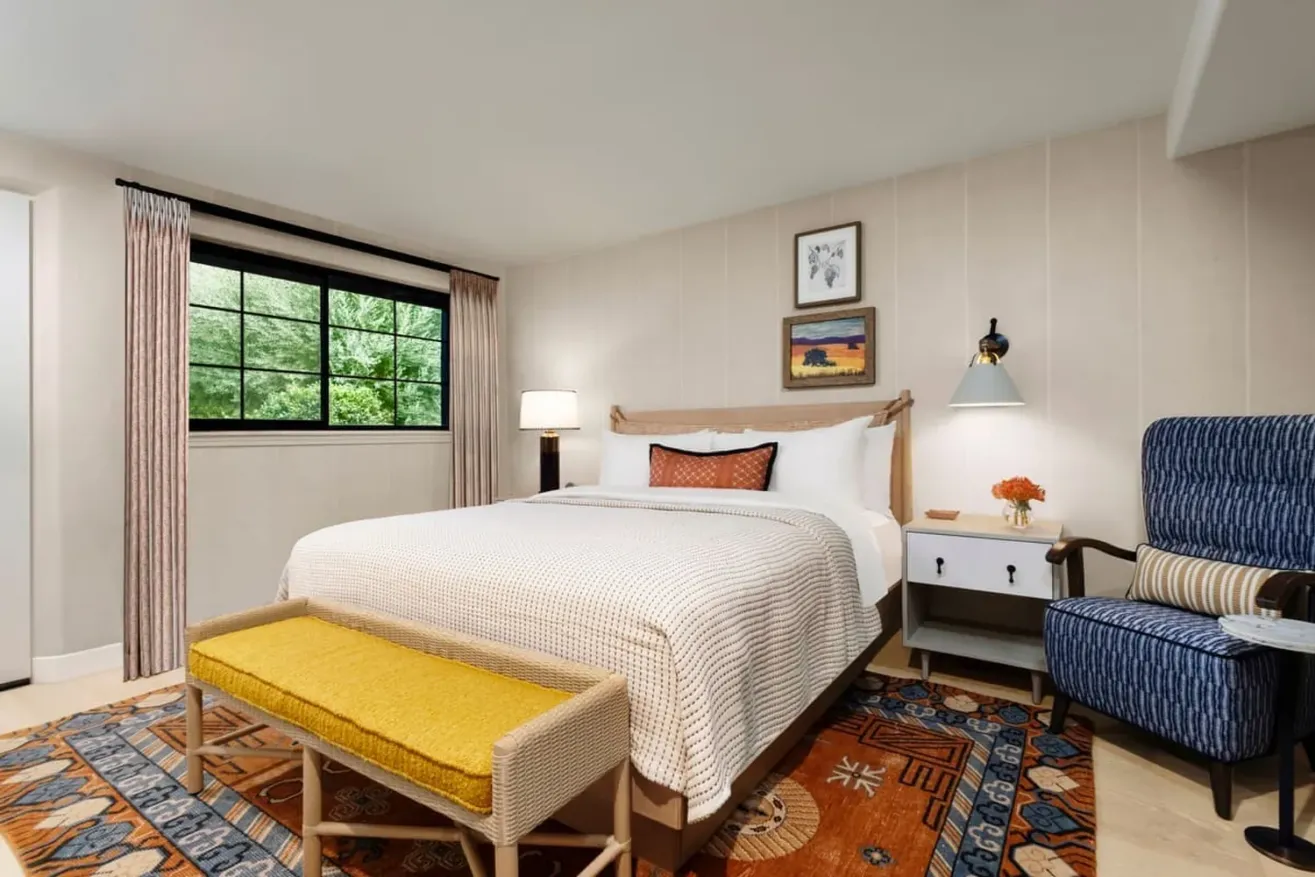 A cozy bedroom with a large bed, patterned rug, yellow bench, armchair, nightstand with lamp, and artwork on light walls. A window with curtains lets in natural light and greenery is visible outside.