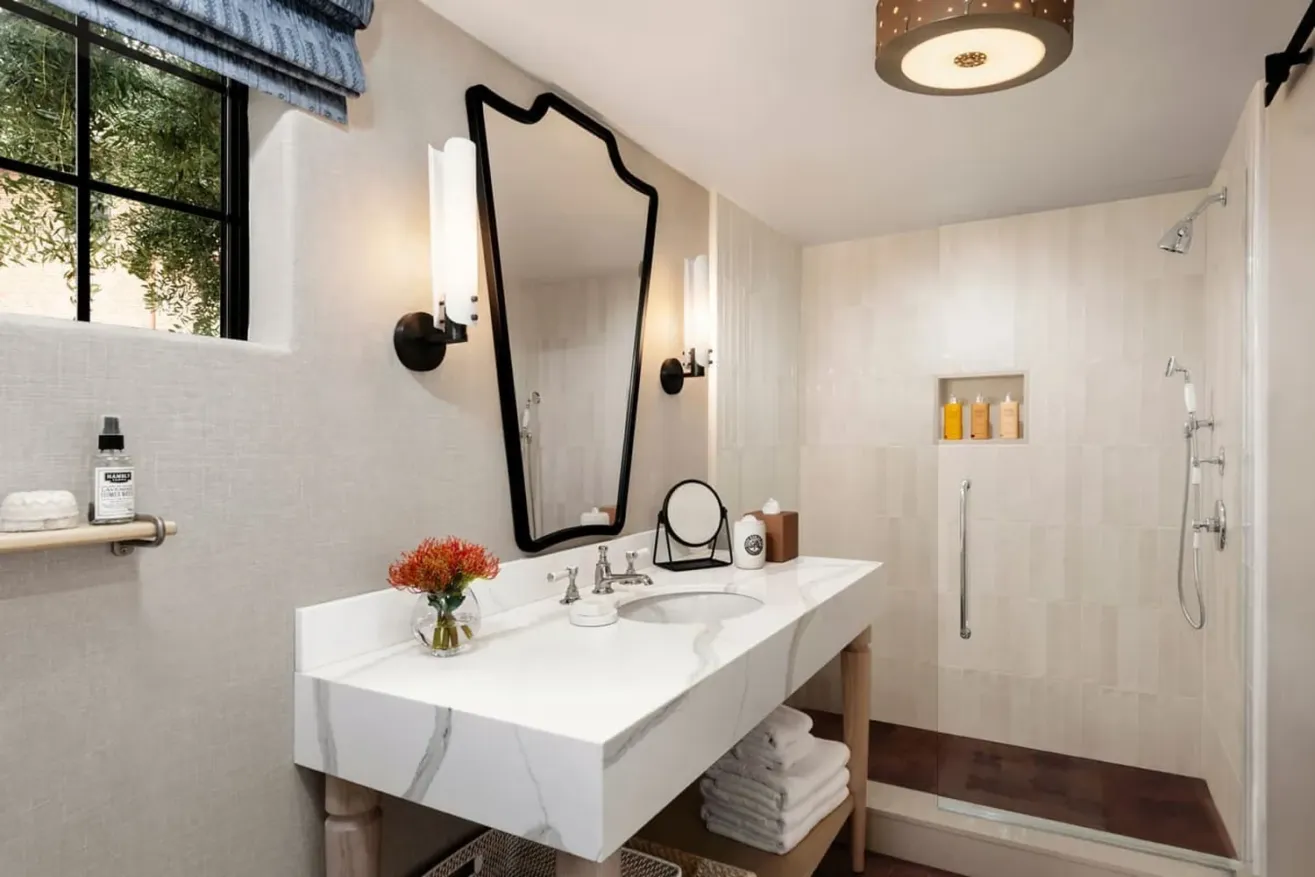 Modern bathroom featuring a white double sink with marble veining, a decorative black-framed mirror, wall sconces, walk-in shower with toiletries, towels on shelves, flowers, and a window with a blue curtain.
