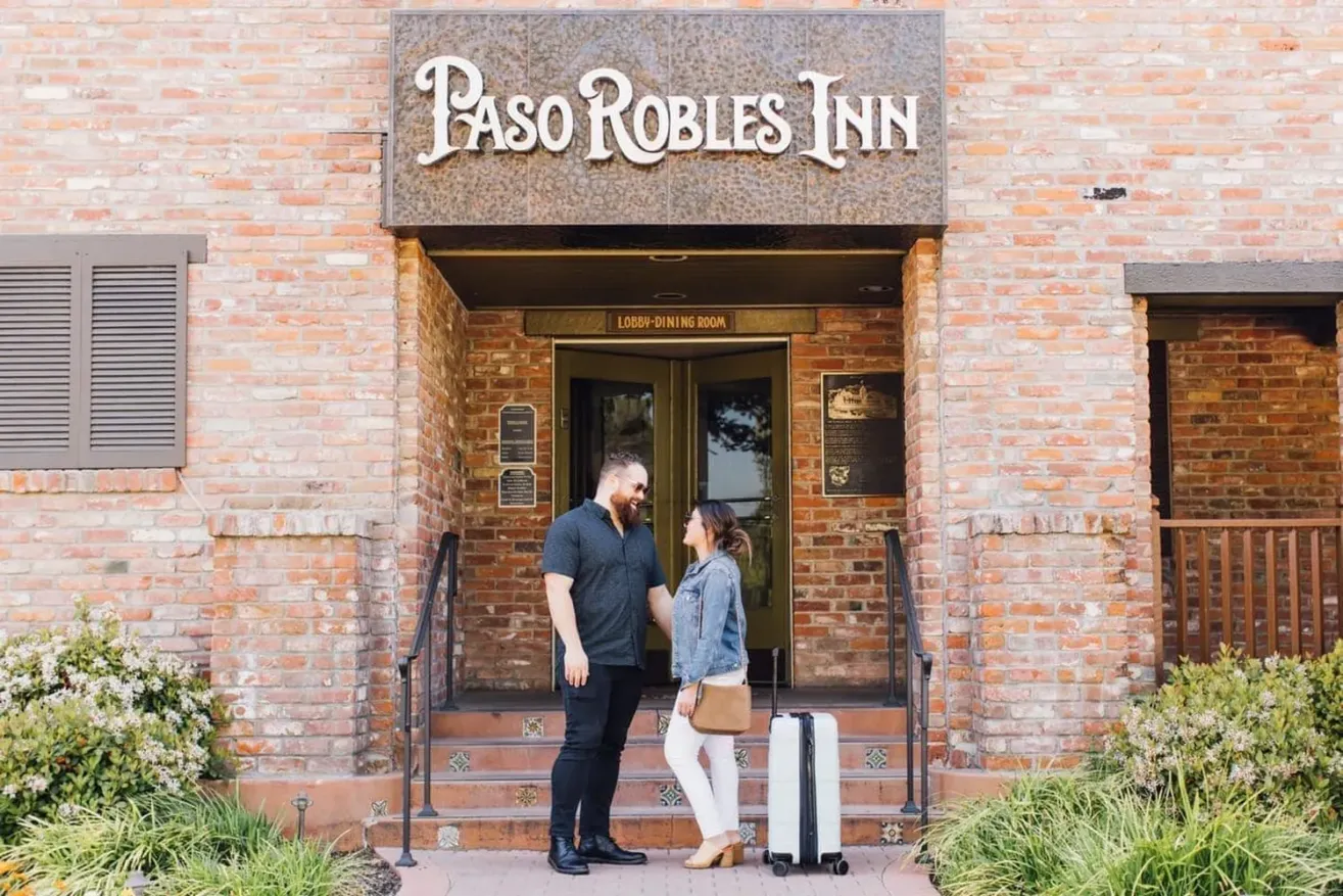 A man and woman stand in front of the Paso Robles Inn entrance, smiling at each other with a white suitcase beside them. The inn has a brick exterior and plants on both sides of the doorway.
