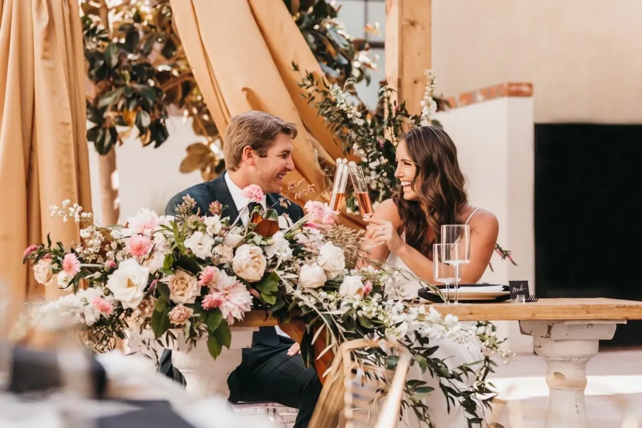 A bride and groom sit at a decorated table outdoors, smiling and clinking glasses. The table is adorned with pink and white flowers, and golden drapery hangs in the background.