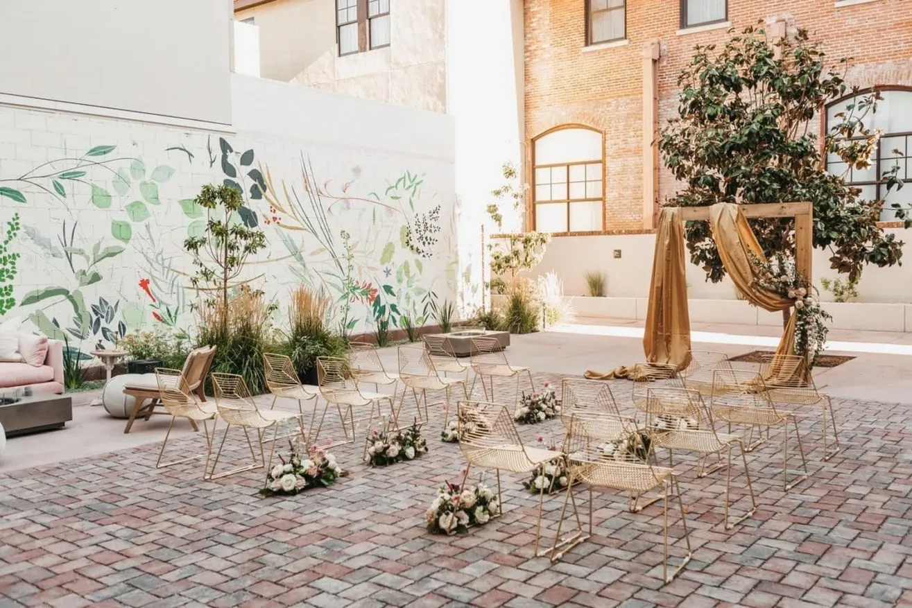 Outdoor wedding ceremony setup with gold chairs arranged in rows, floral arrangements on the ground, a modern geometric arch draped in gold fabric, and a mural of plants on the wall in a courtyard with brick flooring.
