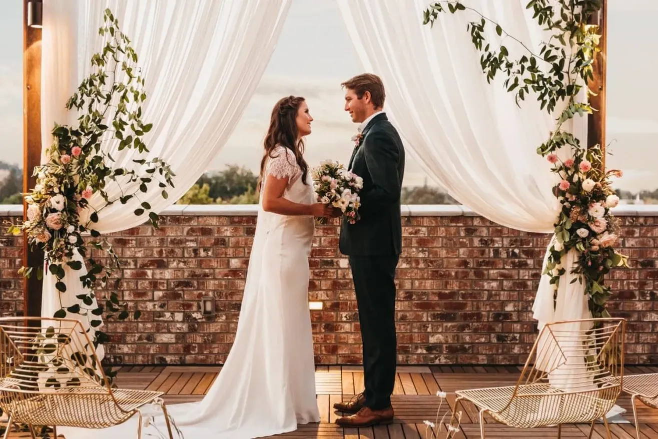 A bride and groom stand facing each other under a floral wedding arch with white drapes, holding hands and smiling, on a wooden deck with metal chairs and a brick wall in the background.