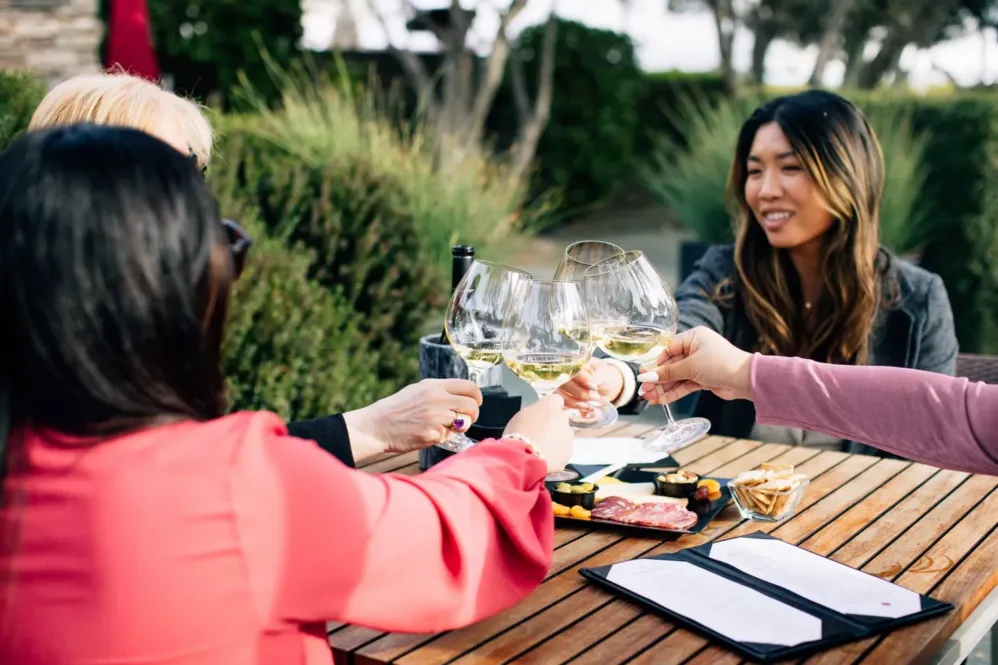 Four women sit around a wooden table outdoors at the Paso Robles Inn, clinking glasses of white wine. With cheese, charcuterie, and a menu on the table, they enjoy cheerful conversation and relaxed Paso Robles Inn Experiences.