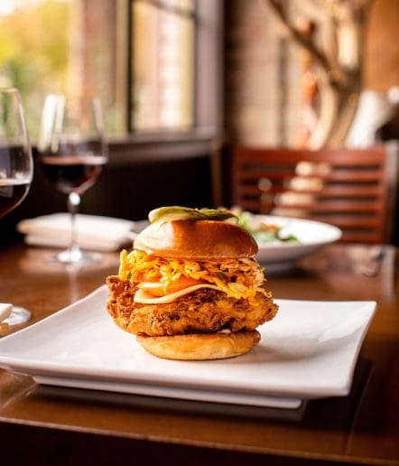 A crispy fried chicken sandwich topped with coleslaw and pickles sits on a square white plate, with two glasses of red wine and a salad in the blurred background at Steakhouse Paso Robles.