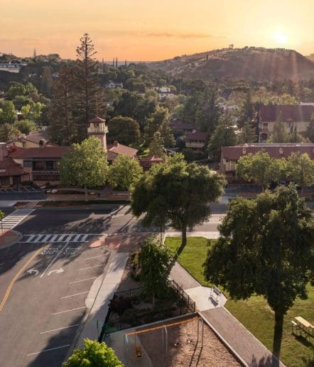 A scenic view of a quiet town at sunset, with tree-lined streets, red-roofed buildings, a playground, and hills in the background under a golden sky.