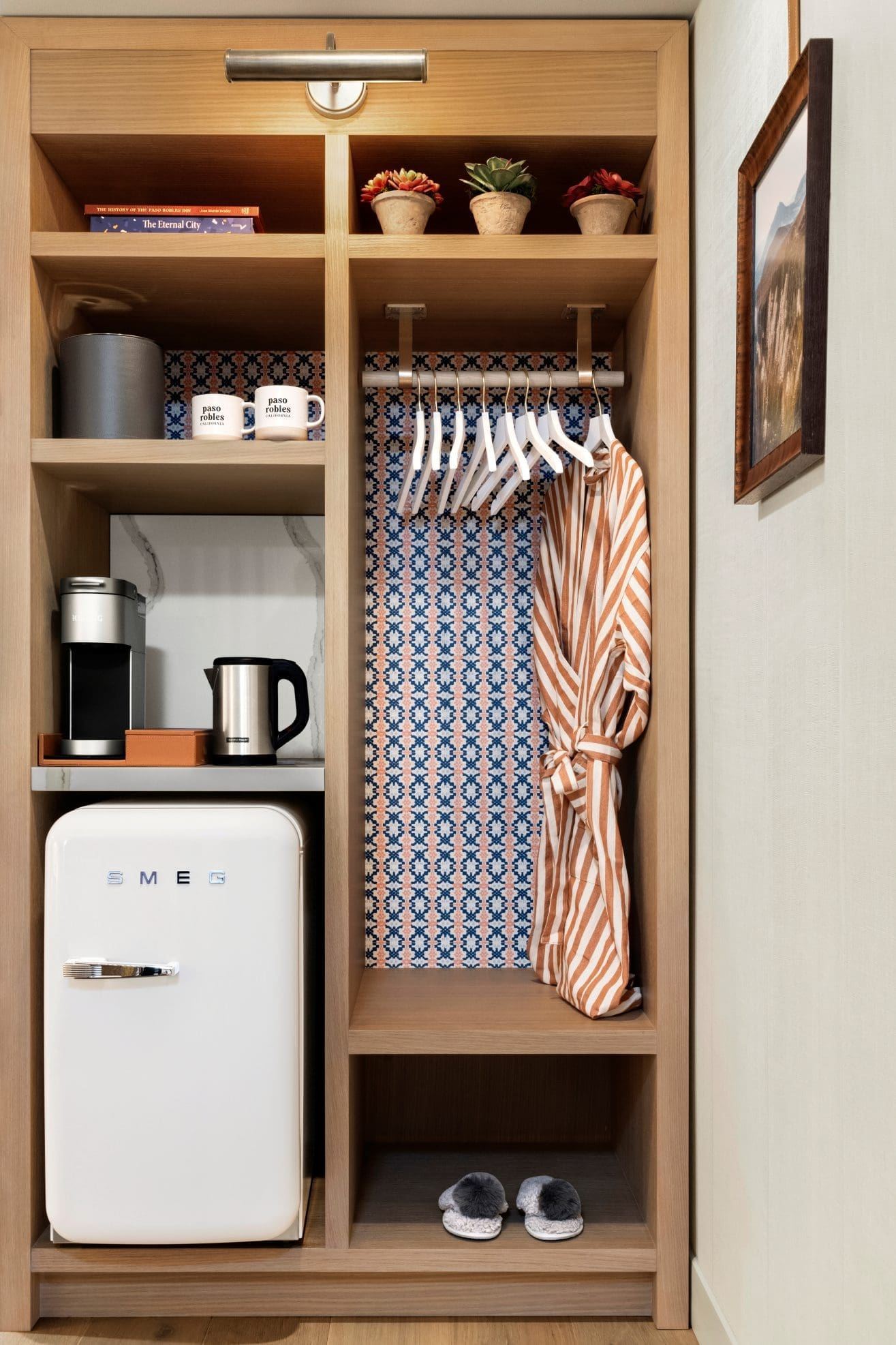 Open wooden wardrobe with a mini SMEG fridge, mugs, electric kettle, striped robe on hangers, potted plants, slippers, and blue patterned wallpaper on the back panel. Framed picture hangs on the wall beside it.