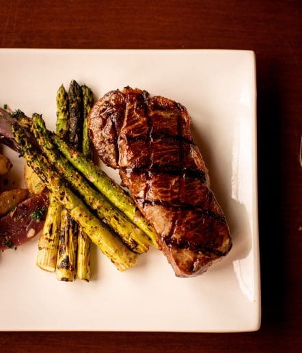 A grilled steak with char marks is served on a white plate alongside roasted asparagus spears and sliced potatoes, evoking the refined flavors of a top Steakhouse Paso Robles, all set on a dark wooden table.