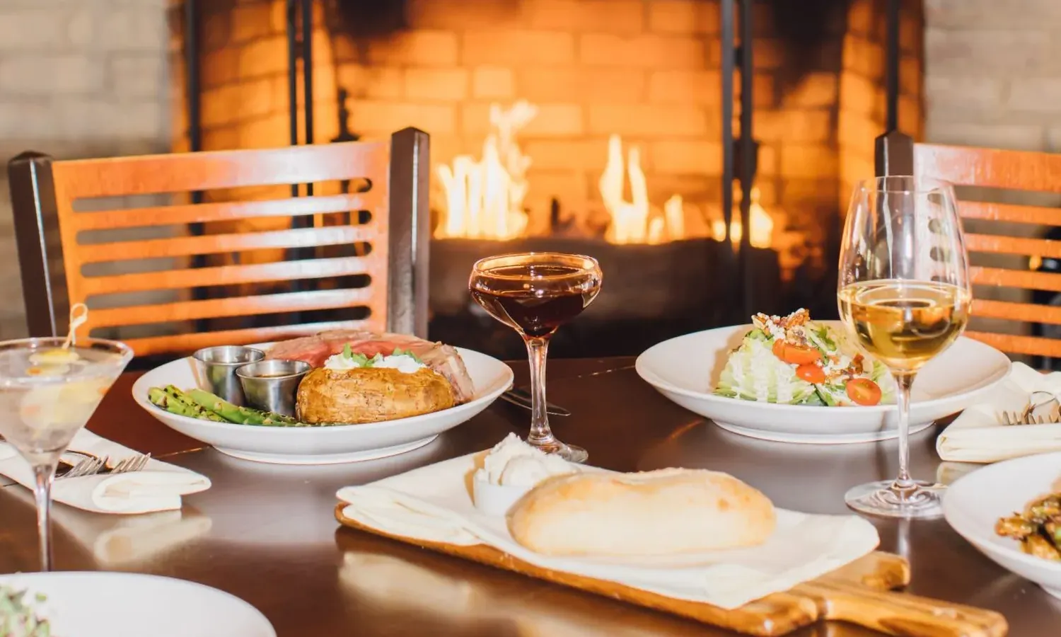 A cozy Steakhouse Paso Robles table set with plates of food, including a baked potato, steak, salad, and bread roll. Two wine glasses and a cocktail complete the scene, with a lit fireplace glowing in the background.