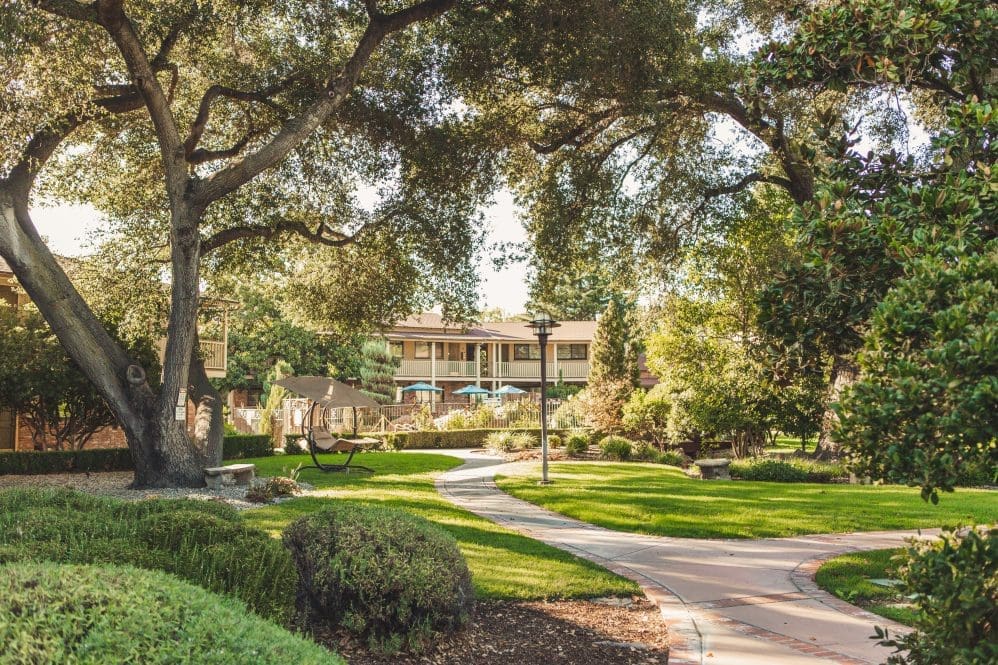 A winding path curves through a lush, green garden with manicured bushes, large trees providing shade, and a building with balconies and outdoor seating in the background.
