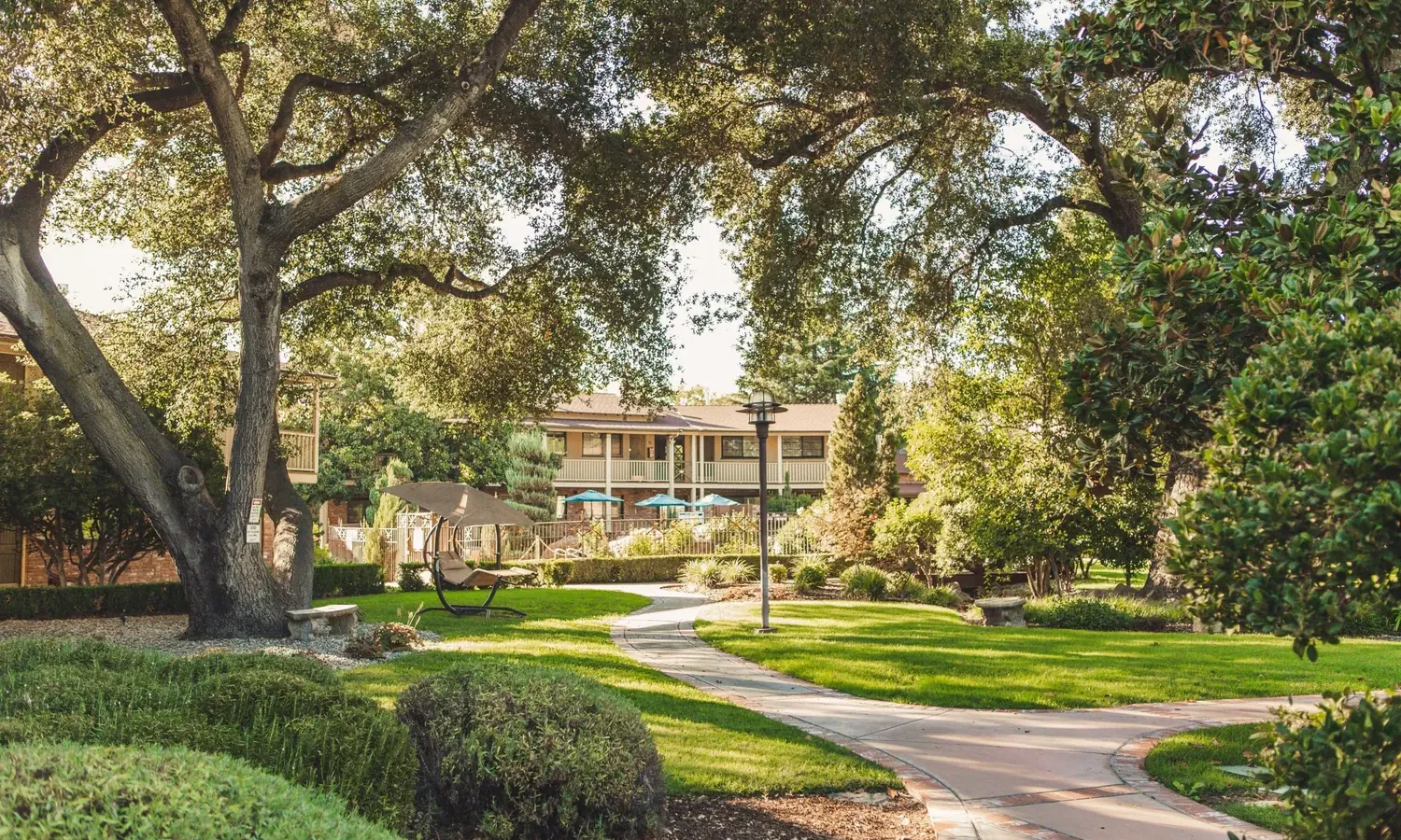 A winding path curves through a lush, green garden with manicured bushes, large trees providing shade, and a building with balconies and outdoor seating in the background.