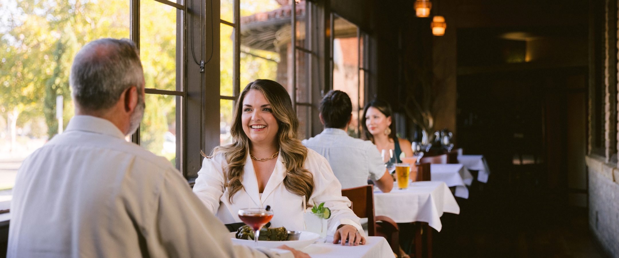 A woman and a man sit at a Steakhouse Paso Robles table by the window, smiling and talking. There are drinks and food on the table. Another couple is seated further down the row, as warm lighting fills the room.