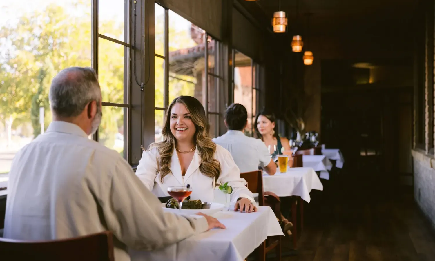 A woman and a man sit at a Steakhouse Paso Robles table by the window, smiling and talking. There are drinks and food on the table. Another couple is seated further down the row, as warm lighting fills the room.