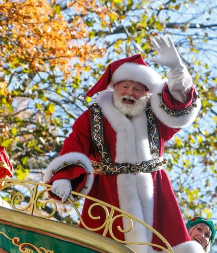 Santa Claus, dressed in a red and white fur-trimmed suit, waves cheerfully from a parade float during Paso Robles Events. Autumn leaves frame the scene, with a festive elf partially visible beside him.
