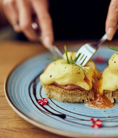 A person uses a knife and fork to cut into eggs Benedict topped with hollandaise sauce and chives, served on a blue plate with decorative sauce and edible flowers.