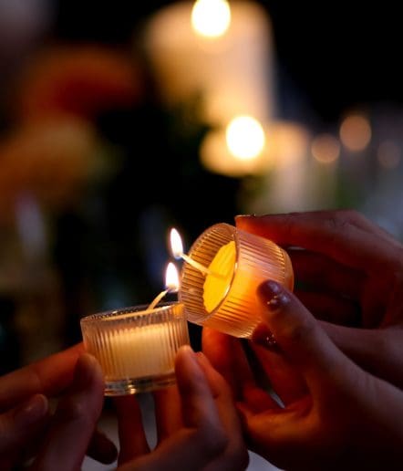Hands hold two small glass candles, one being lit from the flame of another, with blurred glowing candles in the background creating a warm, intimate atmosphere—perfect for memorable Paso Robles Events.