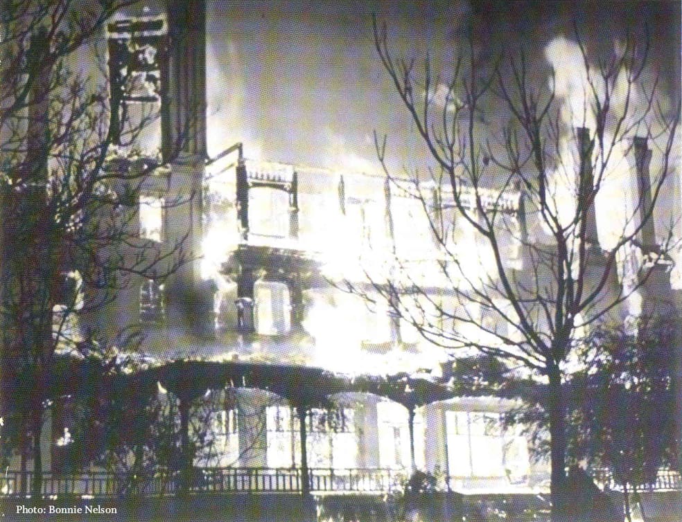 A large building is engulfed in flames at night, with fire consuming the structure and thick smoke rising. Leafless trees are visible in the foreground, silhouetted against the bright fire.