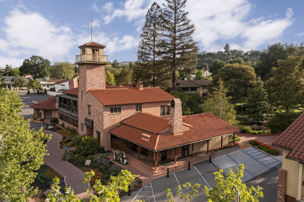 Aerial view of a large brick building with a red-tiled roof and a tall clock tower, surrounded by trees, gardens, parking spaces, and nearby houses under a partly cloudy sky.
