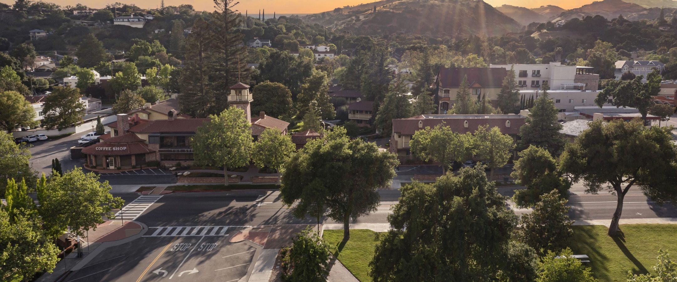 Aerial view of a small town at sunset, featuring tree-lined streets, red-roofed buildings, a coffee shop, a crosswalk, and mountains in the background under a golden sky.
