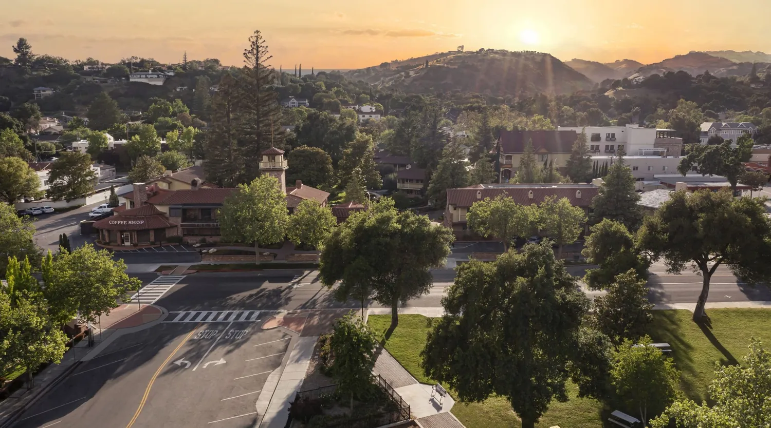 Aerial view of a small town at sunset, featuring tree-lined streets, red-roofed buildings, a coffee shop, a crosswalk, and mountains in the background under a golden sky.
