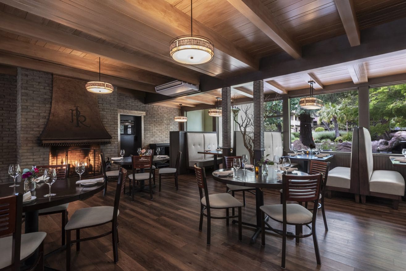 Upscale Steakhouse Paso Robles interior with wooden floors and ceiling, brick fireplace, elegant tables set with glassware, cushioned booths, and large windows overlooking a lush green outdoor area. Warm lighting creates a cozy ambiance.